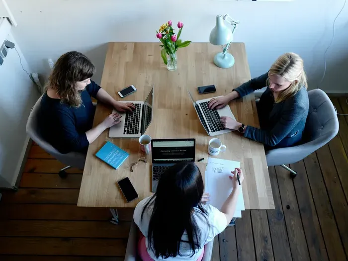 Overhead view of three women working together on laptops and notes at a wooden desk in a modern office on localservicenet.com